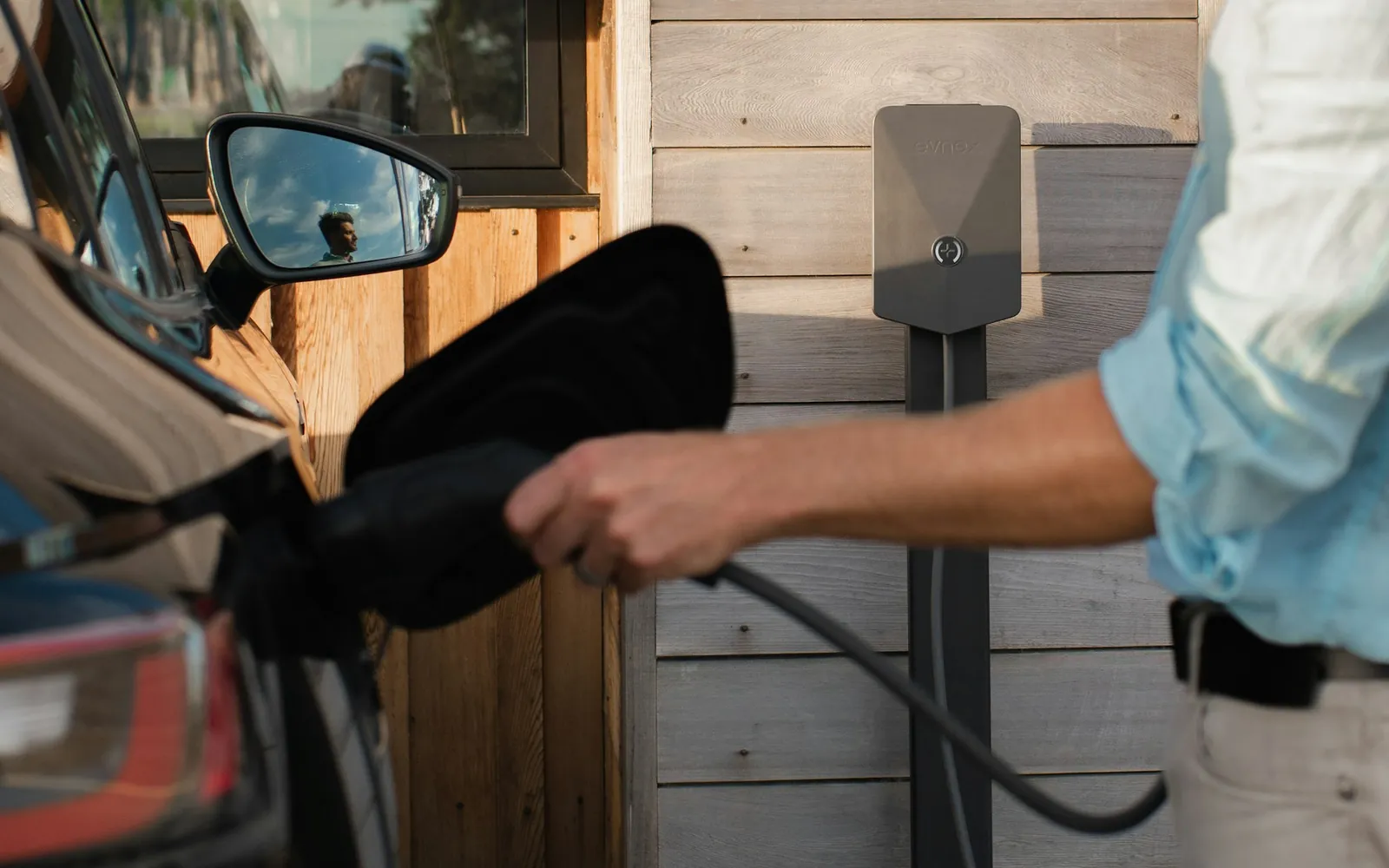 A person holds a charging connector next to an electric car, with a wall-mounted home charger on the wood siding behind.